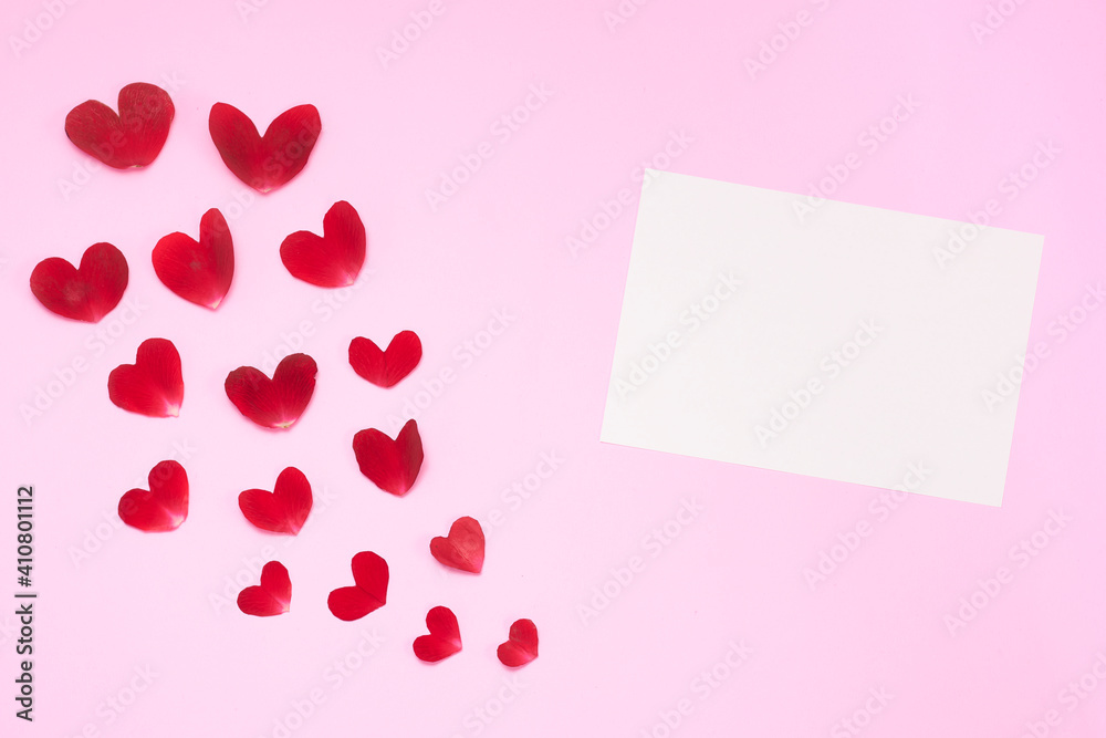Pink background with rose petals in the shape of a heart, with rose, white candles and paper.