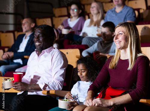 Portrait of laughing european woman watching funny movie in cinema with her african husband and preteen daughter