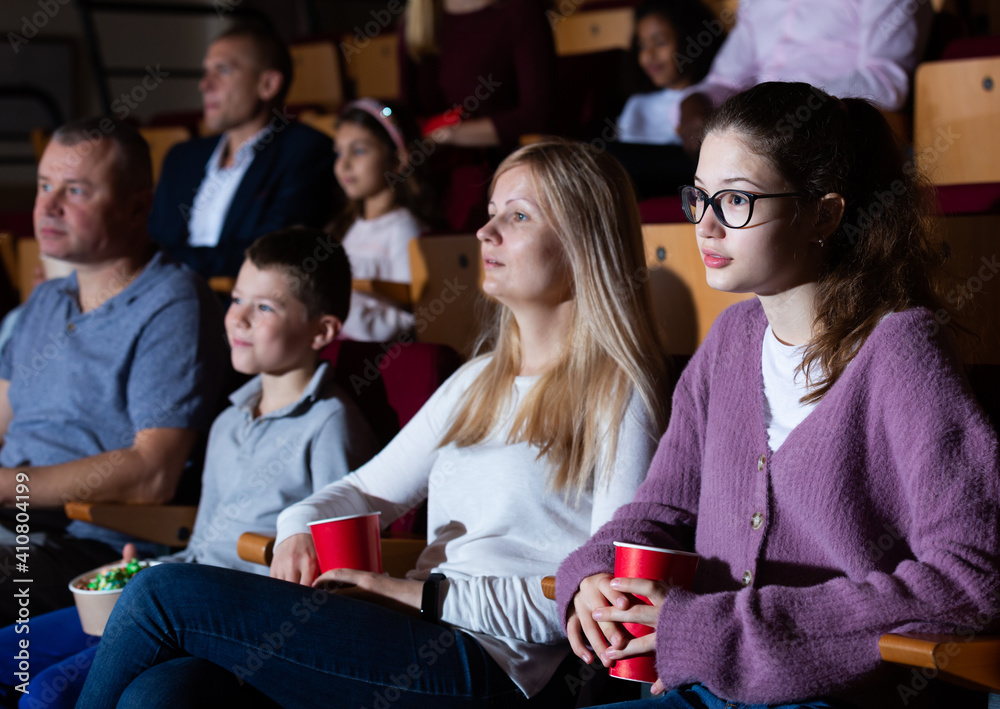Friendly family of four sitting with popcorn and drinks in cinema, watching movie with interest. Family leisure and entertainment concept