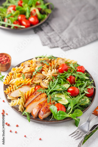 Grilled chicken breast, pasta fusilli, arugula, tomatoes, cucumber slice and mushrooms in plate.