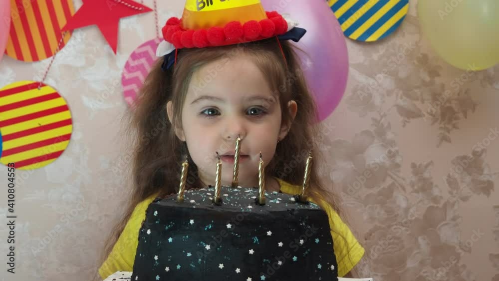 Portrait of little pretty girl with birthday cake a girl in a cap celebrates her birthday, has fun, blows a pipe, blows out the candles on the cake, eats a cake, drinks juice, wears a mask 
