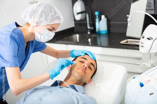Professional beautician performing innovative anti-aging carboxytherapy procedure for facial skin to male client in aesthetic medicine cabinet.