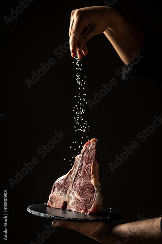 Chef hands cooking dry aged wagyu porterhouse beef steak with large fillet piece adding salt and pepper in a freeze motion on black background. vertical image, place for text photo