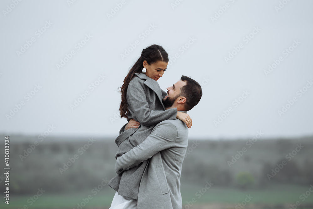 Stylish couple in gray coats on the background of nature. Groom lifts bride up. Happy moments of the wedding day.