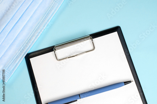A stationery tablet with a pen on a blue background near a medical face mask. Space for text. Medical and business concept.