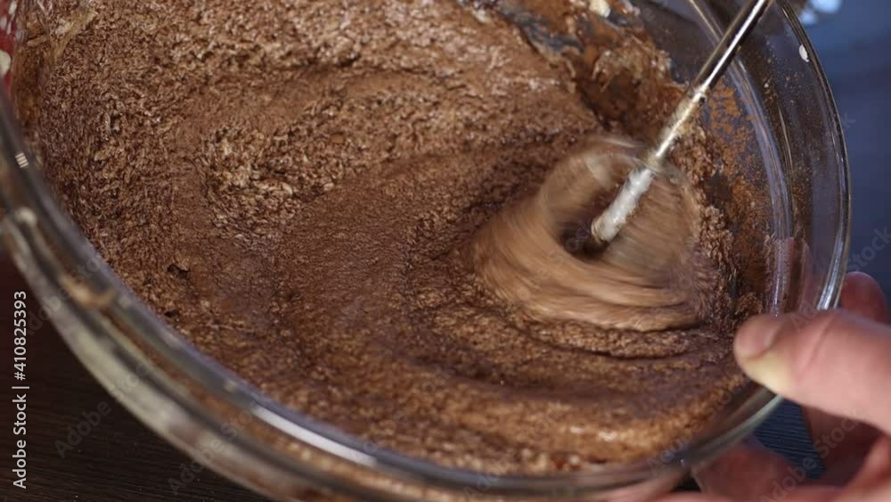 Mixer is mixing chocolate batter for cake in glass bowl. Female is making cupcake