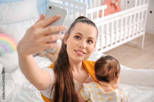 Young mother with little baby in sling taking selfie at home