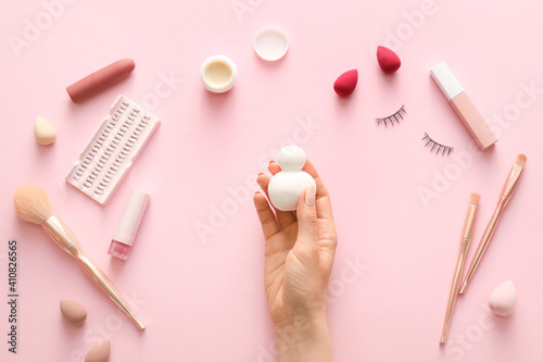 Hand with makeup sponge and cosmetics on color background