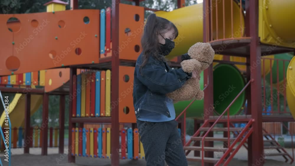 Playful time in epidemic. A happy child spinnig with a teddy bear both in medical mask on the playground.