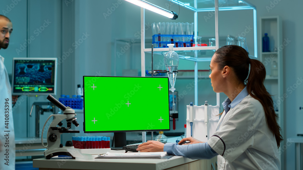 Scientist sitting at desk working on personal computer with mock-up green screen. In background man lab researcher discussing with doctor about vaccine developent bringing blood samples Scientist sitting at desk working on personal computer with mock-up green screen. In background man lab researcher discussing with doctor about vaccine developent bringing blood samples