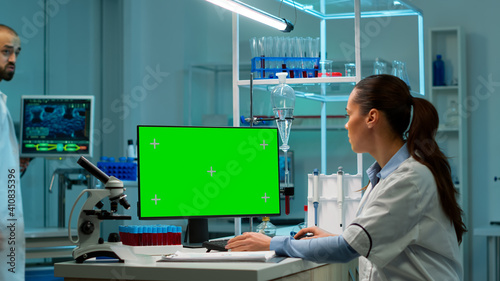Scientist sitting at desk working on personal computer with mock-up green screen. In background man lab researcher discussing with doctor about vaccine developent bringing blood samples