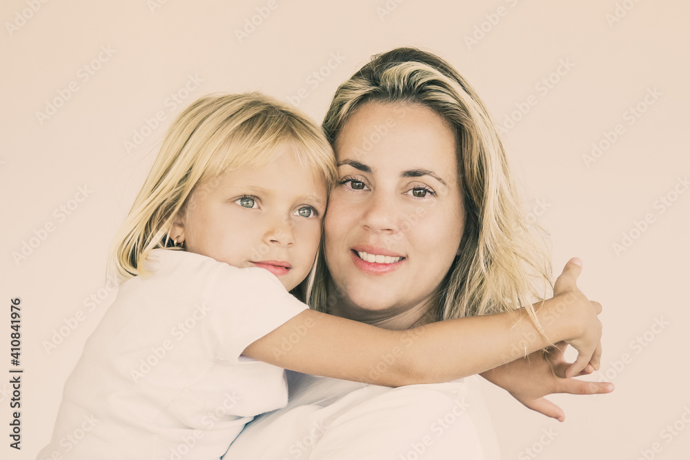 Pretty blonde mother holding daughter and looking at camera. Portrait of adorable little girl sitting on mom hands and embracing or hugging her on white background. Family, love and parenthood concept Pretty blonde mother holding daughter and looking at camera. Portrait of adorable little girl sitting on mom hands and embracing or hugging her on white background. Family, love and parenthood concept