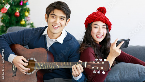 Portrait of cute smiling young Asian lover couple in red and blue long sleeve sweatshirt sitting on the sofa while looking at the camera. Man playing acoustic guitar and woman playing a smartphone