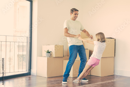 Joyful father playing with daughter among unpacked boxes in new home. Caucasian handsome dad standing in room and holding little girl hands. Family, relocation and moving day concept © Mangostar