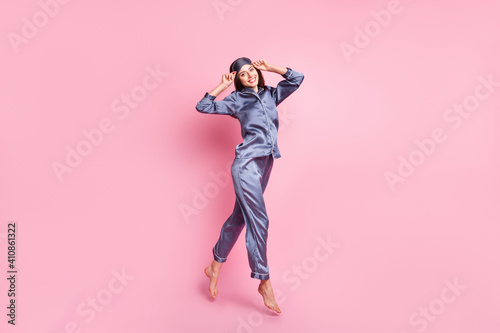 Full length body size photo of brunette in blue pajama jumping smiling touching sleeping mask isolated on pastel pink color background
