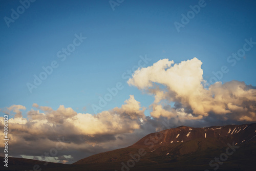 Atmospheric mountain scenery with yellow clouds in blue sky. Scenic landscape with illuminating sunset clouds above mountains with snow. Beautiful sunrise in mountains. Snow on rocks in sunlight.