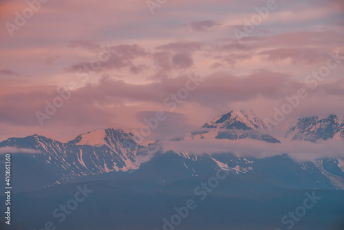 Awesome mountain landscape with great snowy mountain range lit by orange dawn sun among low clouds. Minimal alpine scenery with snowy high mountain ridge under cloudy sky at sunset or at sunrise.