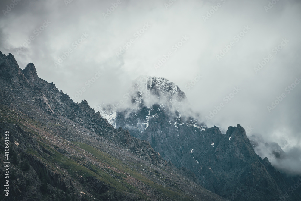 Dark atmospheric surreal landscape with dark rocky mountain top in low clouds in gray cloudy sky. Gray low cloud on high pinnacle. High black rock with snow in low clouds. Surrealist gloomy mountains.