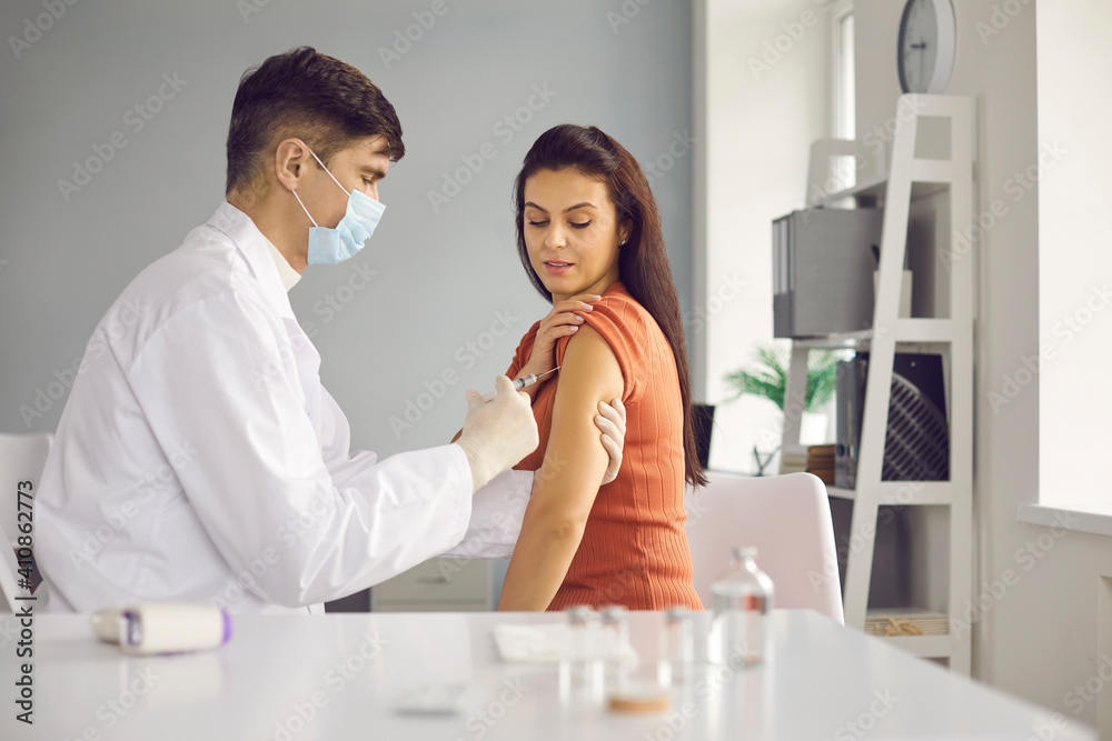 Side view of a male doctor in a medical mask giving a vaccine to a woman for seasonal flu or an outbreak of an infection. Doctor provides services to a patient in a hospital or private clinic. Side view of a male doctor in a medical mask giving a vaccine to a woman for seasonal flu or an outbreak of an infection. Doctor provides services to a patient in a hospital or private clinic.