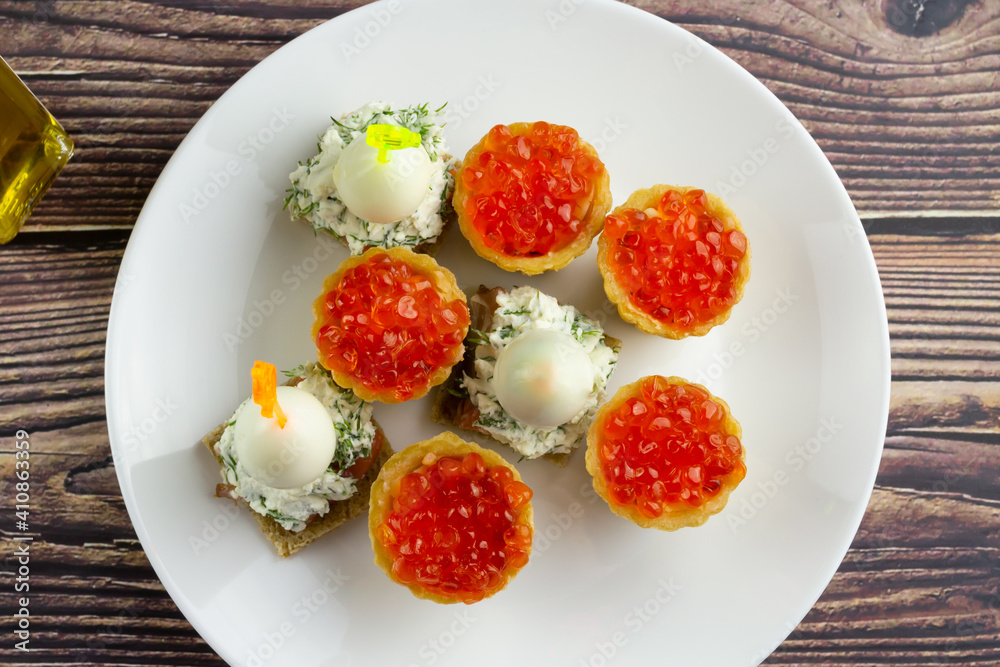 canapes with red caviar, salmon and quail eggs on a white plate, tartlets, small appetizing sandwiches on skewers, bread with cream cheese on a wooden background canapes with red caviar, salmon and quail eggs on a white plate, tartlets, small appetizing sandwiches on skewers, bread with cream cheese on a wooden background