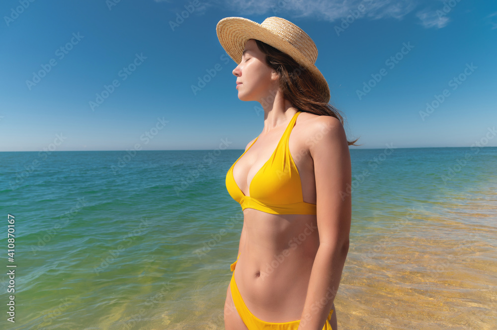 Attractive caucasian young woman in a yellow swimsuit stands on the shore of the azure sea in a straw hat with her eyes closed from pleasure