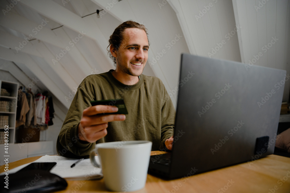 Caucasian male student smiling while sitting at desk successfully affording monthly rent enjoying hot coffee Caucasian male student smiling while sitting at desk successfully affording monthly rent enjoying hot coffee