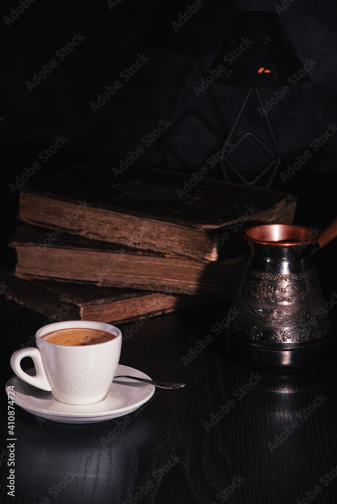White cup of fresh hot coffee, copper cezve and stack of old books on black wooden table in the dark. Low key. Copy space.