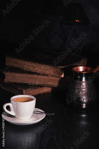 Obraz na płótnie White cup of fresh hot coffee, copper cezve and stack of old books on black wooden table in the dark. Low key. Copy space. w salonie