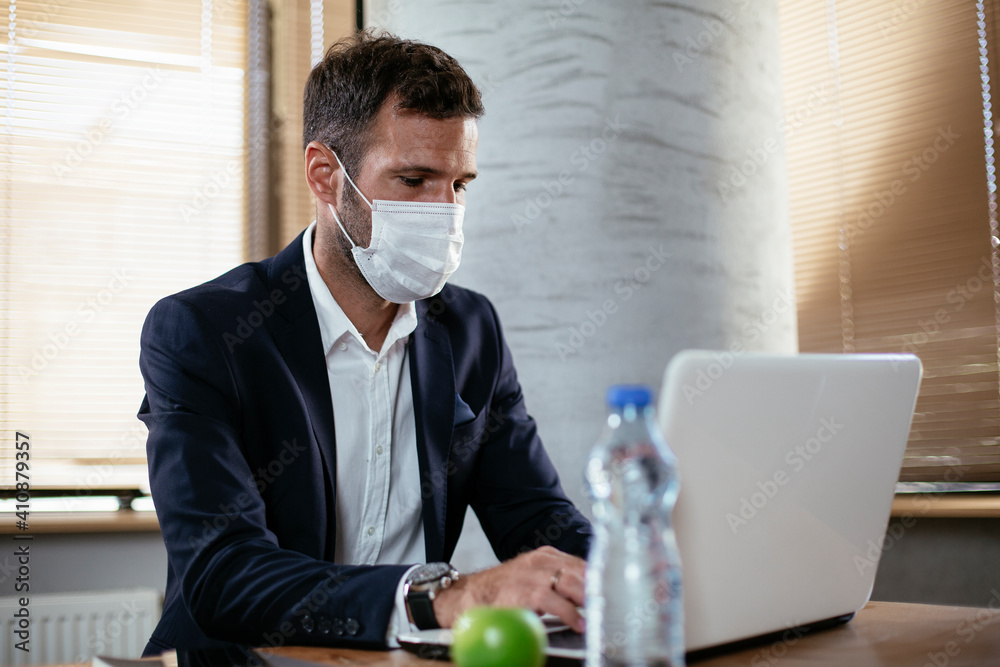 Young businessman with a mask working in his office on laptop. COVID - 19 virus protection.. Young businessman with a mask working in his office on laptop. COVID - 19 virus protection..