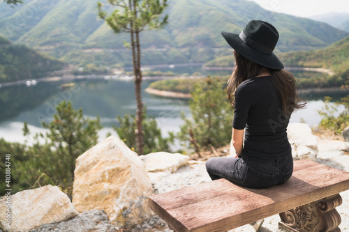 Lonely woman with black hat and black clothes sitting on wooden bench with mountain lake view © mihail