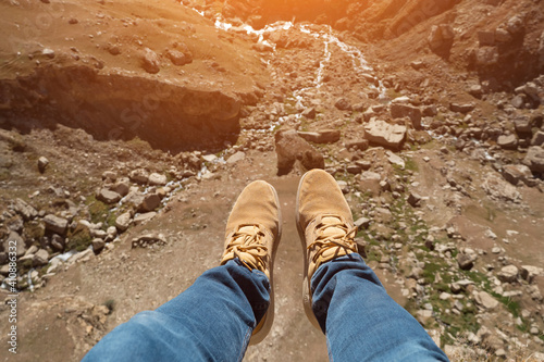 Man's legs over a cliff and a mountain river, sunlight.