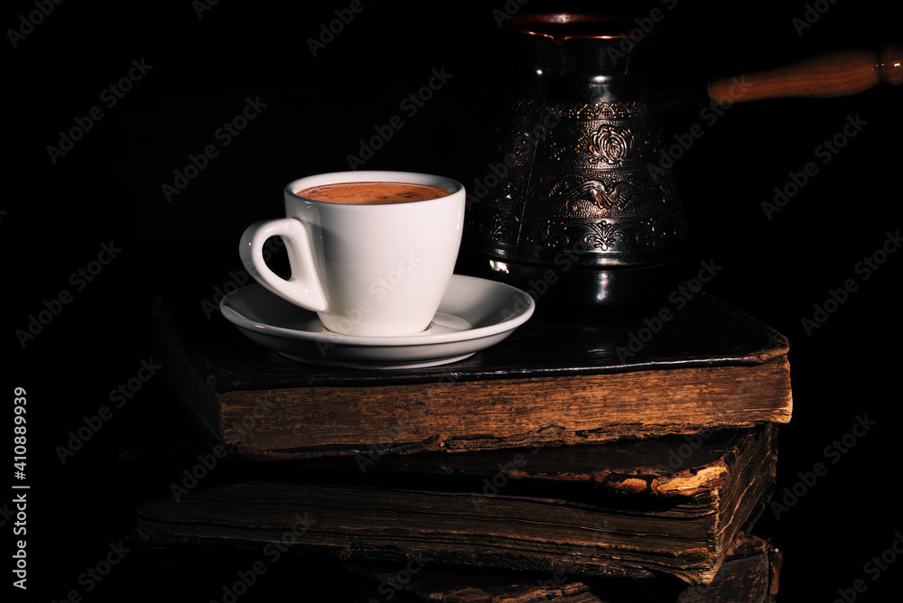 White cup of coffee and copper cezve on stack of old shabby books in the dark. Low key. Closeup.