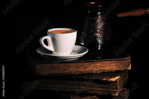 Obraz na płótnie White cup of coffee and copper cezve on stack of old shabby books in the dark. Low key. Closeup. w salonie