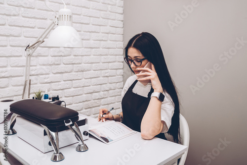 Businesswoman, conducts an appointment for a manicure procedure in a beauty salon. Business lady takes a call from clients. A manicurist in black uniform sits at his desk and speaks on the phone.