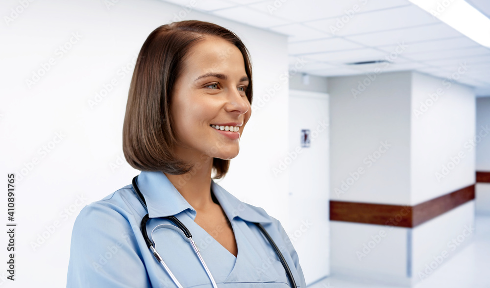 healthcare, medicine and profession concept - smiling female doctor or nurse in blue uniform with stethoscope over hospital corridor background