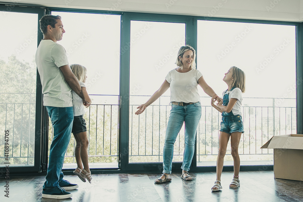 Fototapeta premium Mother, father and two daughters standing in empty living room with open balcony. Happy family relocating in new apartment or house together and watching room. Relocation and moving day concept