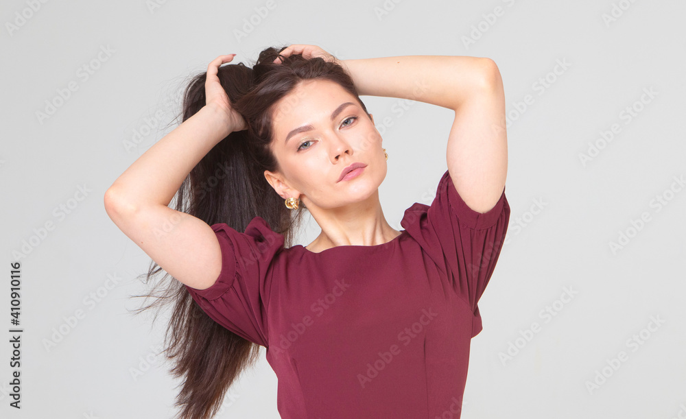 beautiful girl 25 years old, European appearance, with dark long hair, in a red dress, straightens her hair, stands on a light gray background, in the studio. Portrait of a girl, brunette. beauty beautiful girl 25 years old, European appearance, with dark long hair, in a red dress, straightens her hair, stands on a light gray background, in the studio. Portrait of a girl, brunette. beauty