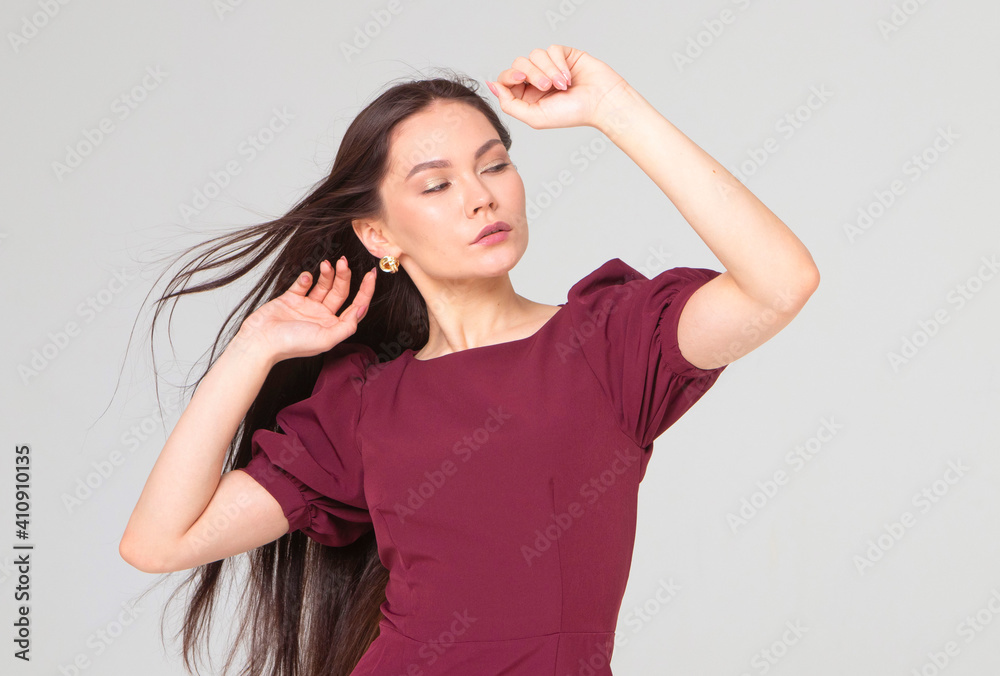 Fototapeta premium beautiful girl 25 years old, European appearance, with dark long hair, in a red dress, straightens her hair, stands on a light gray background, in the studio. Portrait of a girl, brunette. beauty