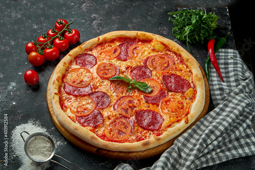 Oven-baked Italian pizza with red sauce, cheese, tomatoes and pepperoni salami in a composition with ingredients on a dark background. View from above.