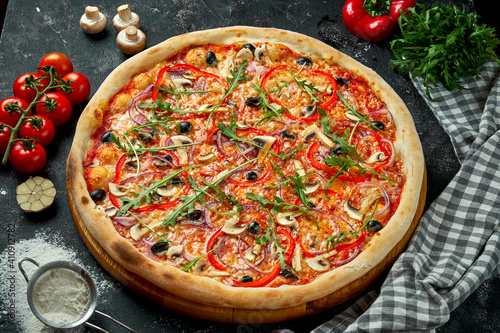 Oven-baked Italian pizza with sauce, cheese, mushrooms, bell peppers and olives in a composition with ingredients on a dark background. Top view. Vegetarian pizza