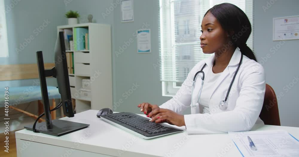 Medium shot of beautiful young african american woman doctor assistant working in medical office using computer, looking in camera smiling. Modern healthcare, online medical help concept Medium shot of beautiful young african american woman doctor assistant working in medical office using computer, looking in camera smiling. Modern healthcare, online medical help concept