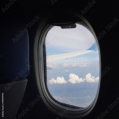 Outlook through plane window while traveling with window surround framing blue sky and clouds with focus on wing outside.