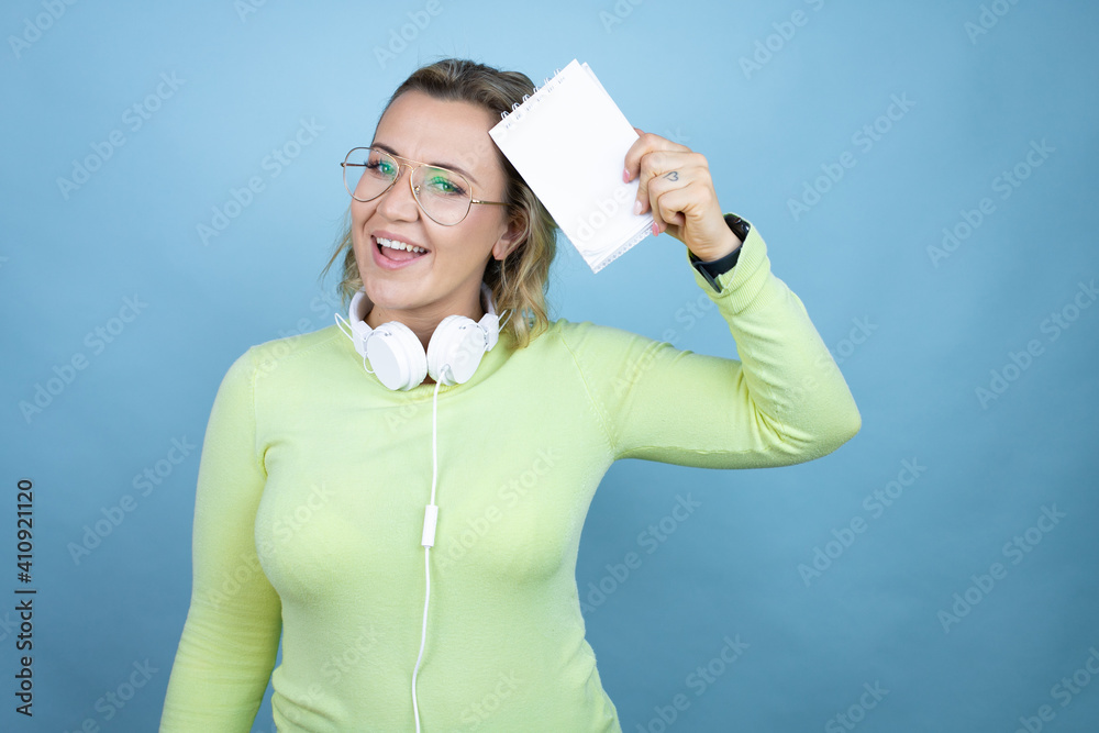 Young caucasian woman wearing headphones on neck over blue background smiling and showing blank notebook in her hand Young caucasian woman wearing headphones on neck over blue background smiling and showing blank notebook in her hand