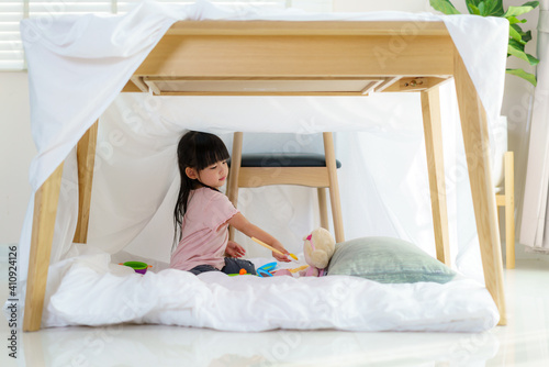 Asian cute little girl play cooking and feeding food to her doll while sitting in a blanket fort in living room at home © ake1150