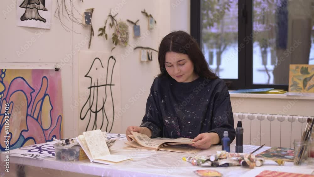 Woman smiling happy reading book in art studio, one young female in the light modern studio with window. Woman in art class with book in hands Woman smiling happy reading book in art studio, one young female in the light modern studio with window. Woman in art class with book in hands