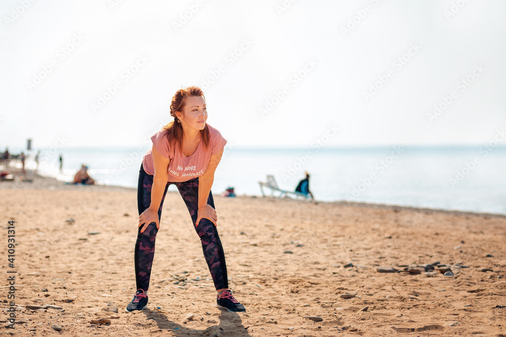 An adult tired beautiful woman in sportswear takes a break during a workout. In the background, the ocean and the beach. Copy space. Outdoor