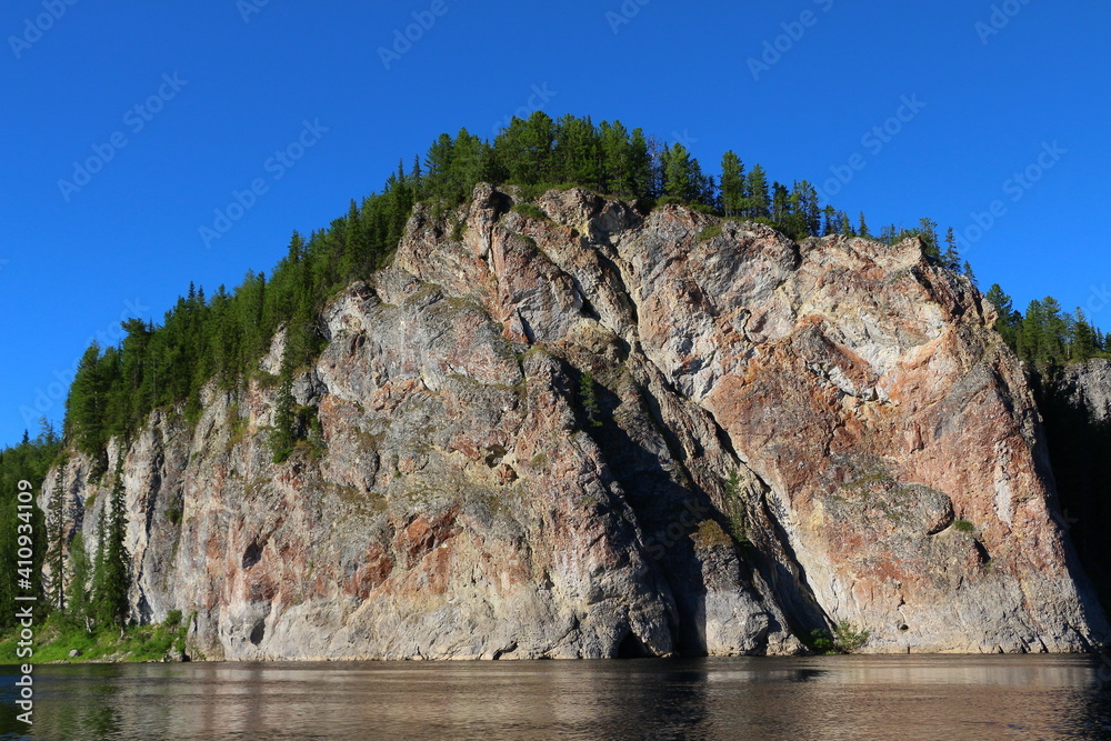 View from the lake on a high steep bald rock with a forest at the top and a cave at the base.Natural mountain landscape on a sunny summer day with a beautiful blue sky View from the lake on a high steep bald rock with a forest at the top and a cave at the base.Natural mountain landscape on a sunny summer day with a beautiful blue sky