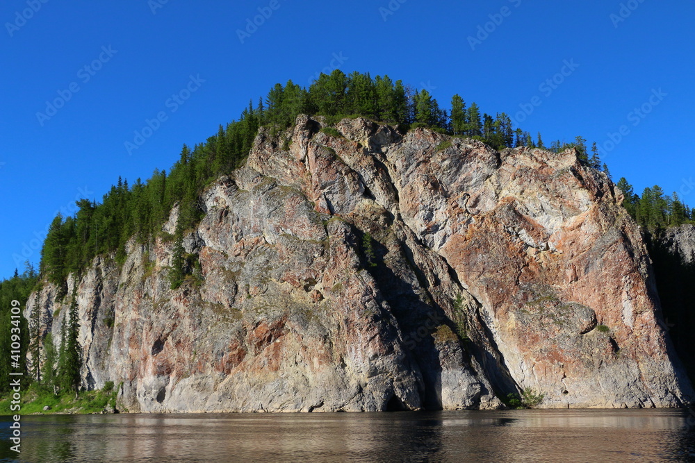 View from the lake on a high steep bald rock with a forest at the top and a cave at the base.Natural mountain landscape on a sunny summer day with a beautiful blue sky