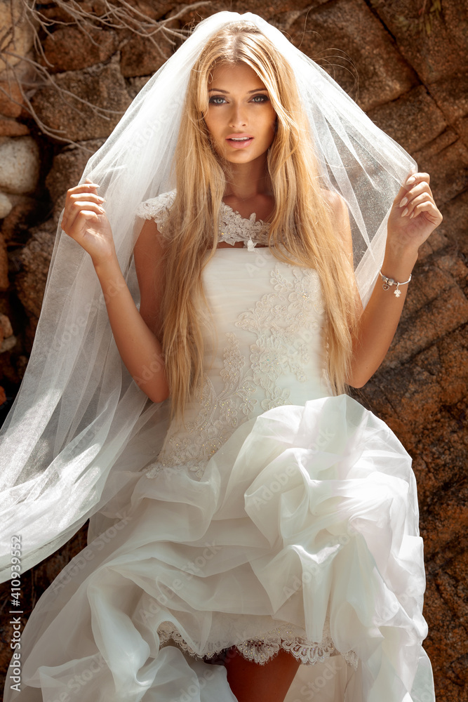Beautiful sexy blonde woman in wedding dress with white veil and long hair and tanned body posing at the stone.