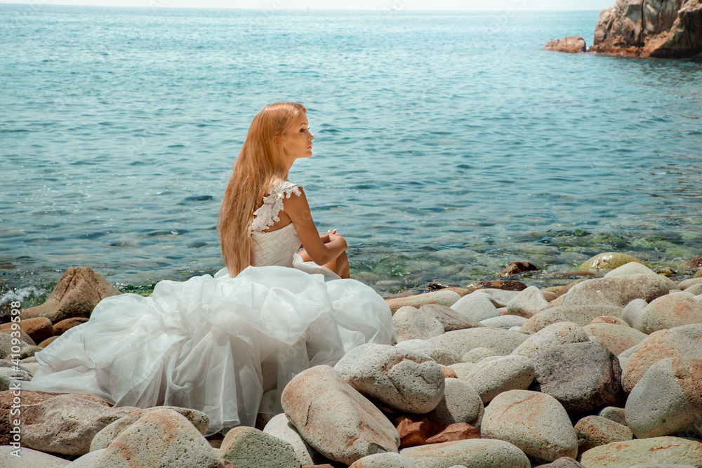 Beautiful sexy blonde woman in wedding dress with long hair and tanned body sitting at the seashore.